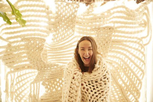 Cheerful woman wrapped in chunky knit blanket smiles in front of macrame wall hanging in a brightly lit indoor setting.