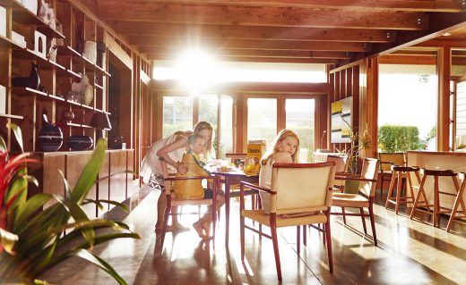 Indoor shot of a woman and two children seated around a table with a box of cereal, bathed in natural light.