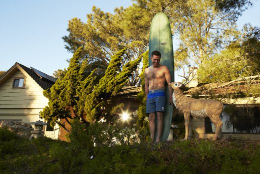 Shirtless man stands with surfboard and deer statue in front of house and trees on a sunny day. Outdoor lifestyle portrait.