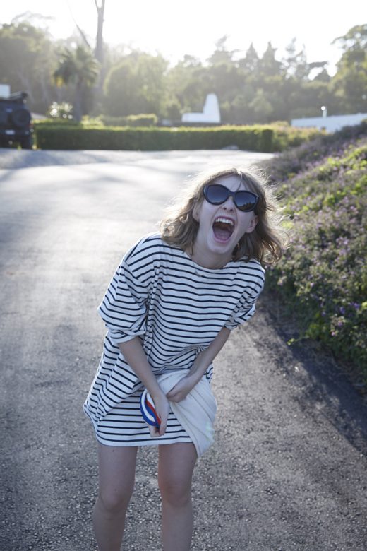 A joyful young woman wearing sunglasses and a striped shirt dress stands on a road, smiling with an open mouth, with a background of trees.
