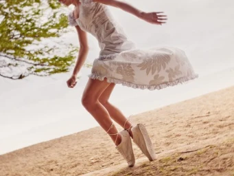 Model in patterned white dress and sneakers jumping on a sandy beach captured by Dewey Nicks.