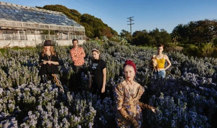 Group of five people standing among purple wildflowers in front of a glass greenhouse under a clear sky