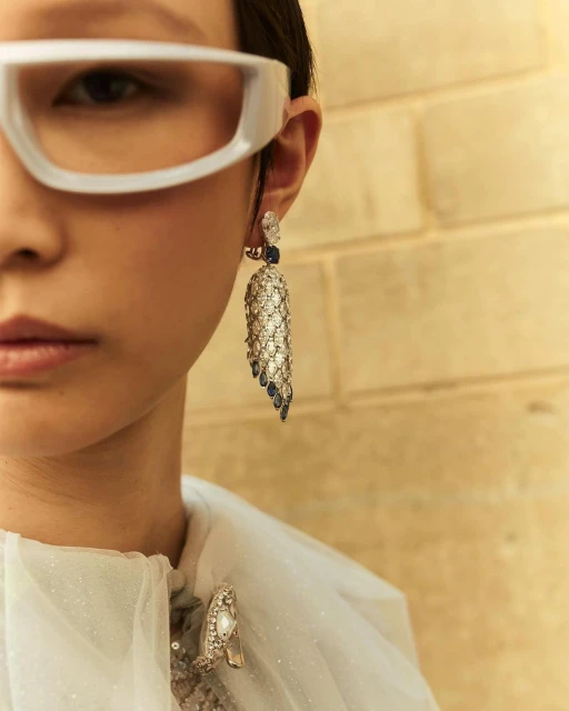 Close-up of an adult female model wearing white-rimmed glasses, an ornate crystal and blue earring, and a decorated sheer top in a studio setting.