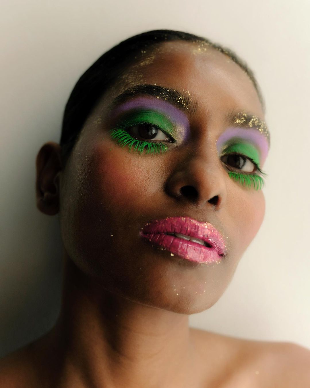 Close-up studio shot of a dark-skinned model wearing vibrant purple, green, and pink makeup with gold glitter accents and false eyelashes.