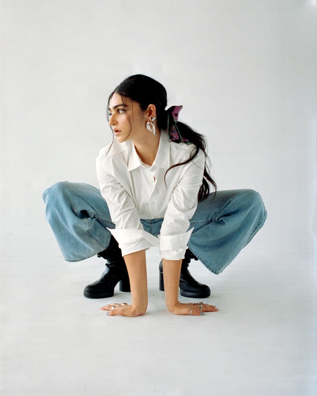 Woman in white shirt and blue jeans squatting against a white background, photographed by Dhruvin