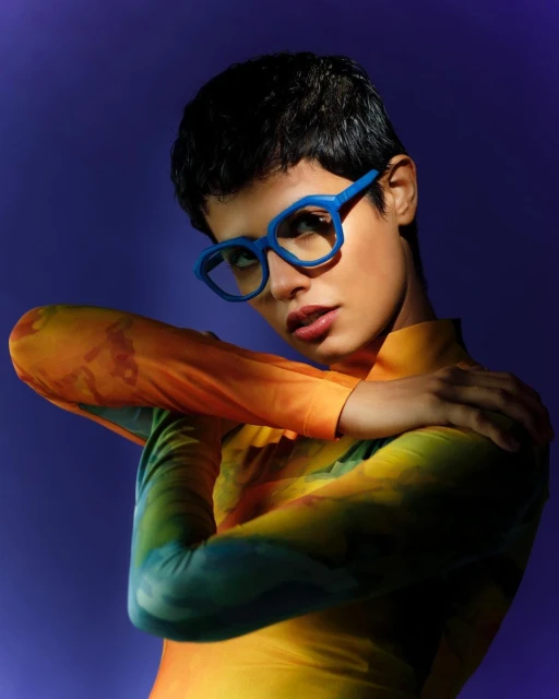 Stylish woman with short hair and blue eyeglasses poses against a purple backdrop, wearing a tie-dye long-sleeve top in studio lighting.