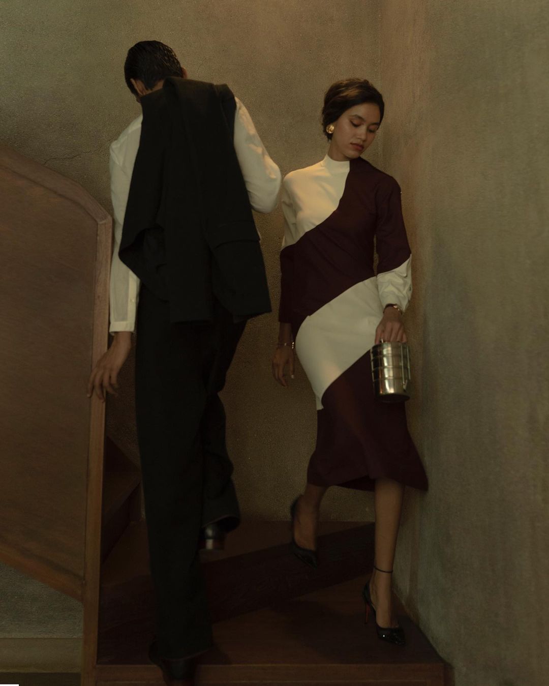 A woman in a two-tone dress walks down wooden stairs carrying a metal purse while a man walks ahead of her.