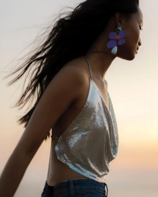 Woman in chainmail top and jeans with oversized flower earrings poses outside against light sky and blowing hair in fashion shot.