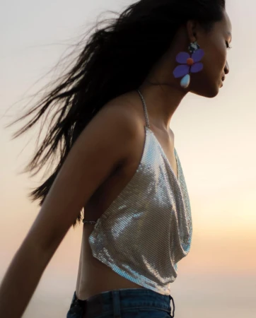 Woman in chainmail top and jeans with oversized flower earrings poses outside against light sky and blowing hair in fashion shot.
