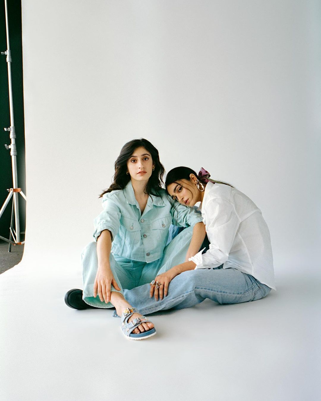 Two young women in casual fashion sitting indoors on a seamless background in a photo studio with soft, natural-looking light.