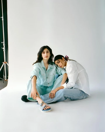 Two young women in casual fashion sitting indoors on a seamless background in a photo studio with soft, natural-looking light.