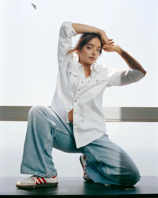 A young woman with long brown hair poses in front of a window, wearing a white button-down shirt, loose jeans, and Adidas sneakers indoors.