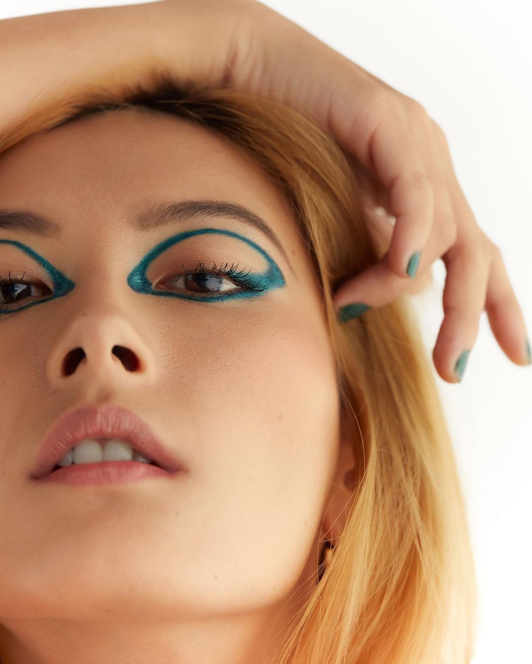 Close-up portrait of a woman with green eyeliner, blonde hair, and green nail polish against a plain white backdrop in a beauty style.