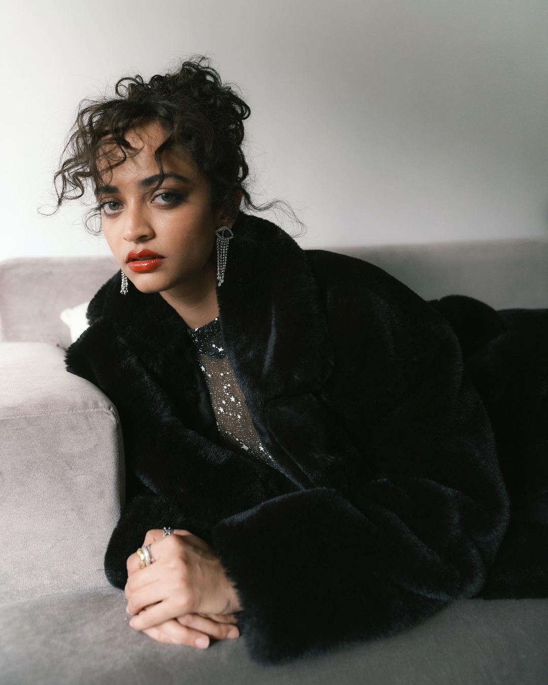 Woman in a black faux fur coat and sparkly top leaning on a grey sofa in Mumbai photographed by Dhruvin.