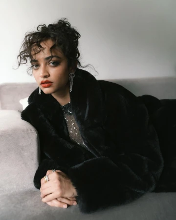 Woman in a black faux fur coat and sparkly top leaning on a grey sofa in Mumbai photographed by Dhruvin.