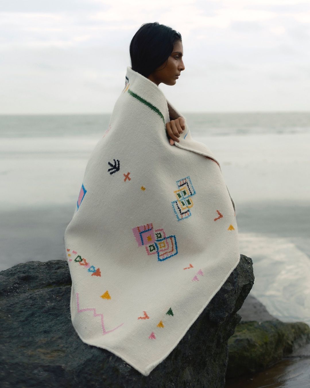 Woman wrapped in embroidered blanket sits on rock at beach with neutral sky and water background, portrait view.