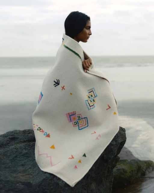 Woman wrapped in embroidered blanket sits on rock at beach with neutral sky and water background, portrait view.