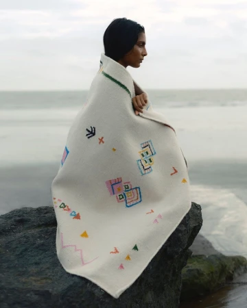 Woman wrapped in embroidered blanket sits on rock at beach with neutral sky and water background, portrait view.