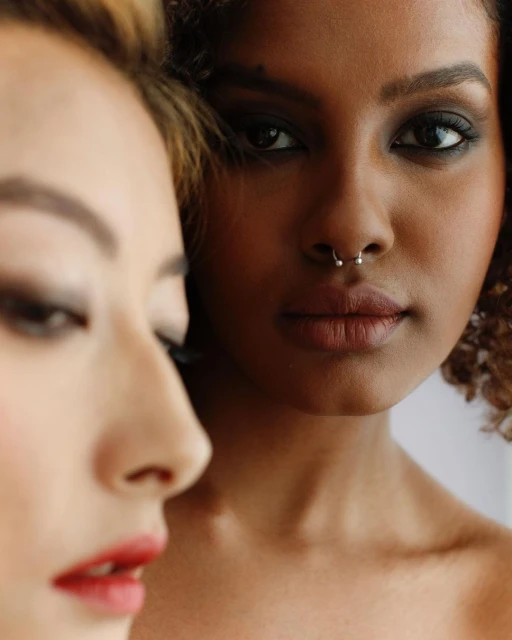 Close-up studio shot of two women with professional makeup, one Asian with red lipstick and one Black with a nose ring.