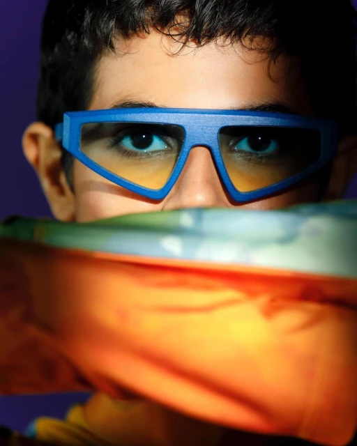 Portrait of a young adult with blue geometric sunglasses and a colorful cloth covering part of their face in front of a purple backdrop.