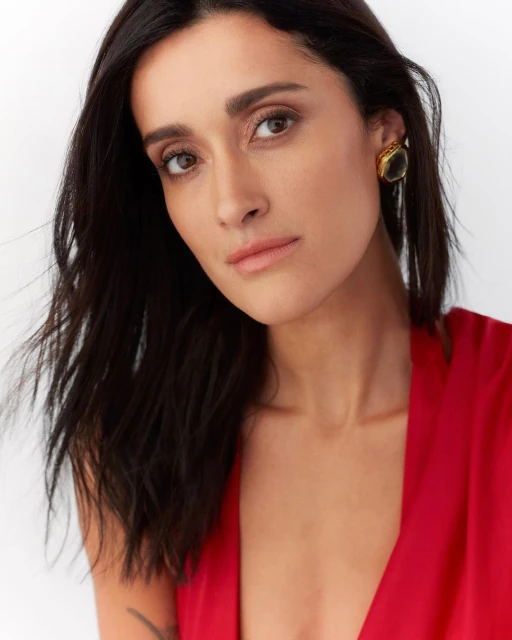 Close-up portrait of a brunette woman with fair skin wearing a red dress and gold earrings against a clean white backdrop.