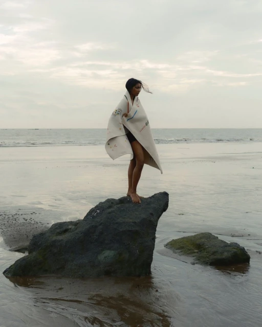 Woman stands on a rock at the beach wrapped in a patterned cloth, looking out at the ocean under a cloudy sky at dusk.