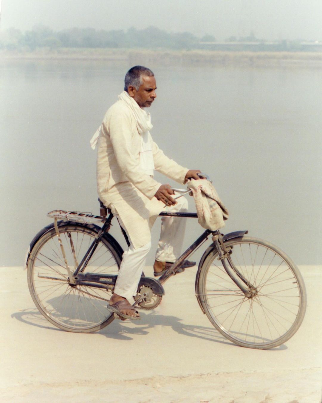 Elderly Indian man in white riding a vintage bicycle on a light-colored road with a river in the background on a cloudy day.