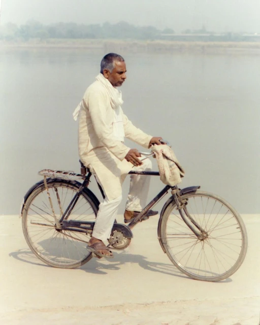 Elderly Indian man in white riding a vintage bicycle on a light-colored road with a river in the background on a cloudy day.