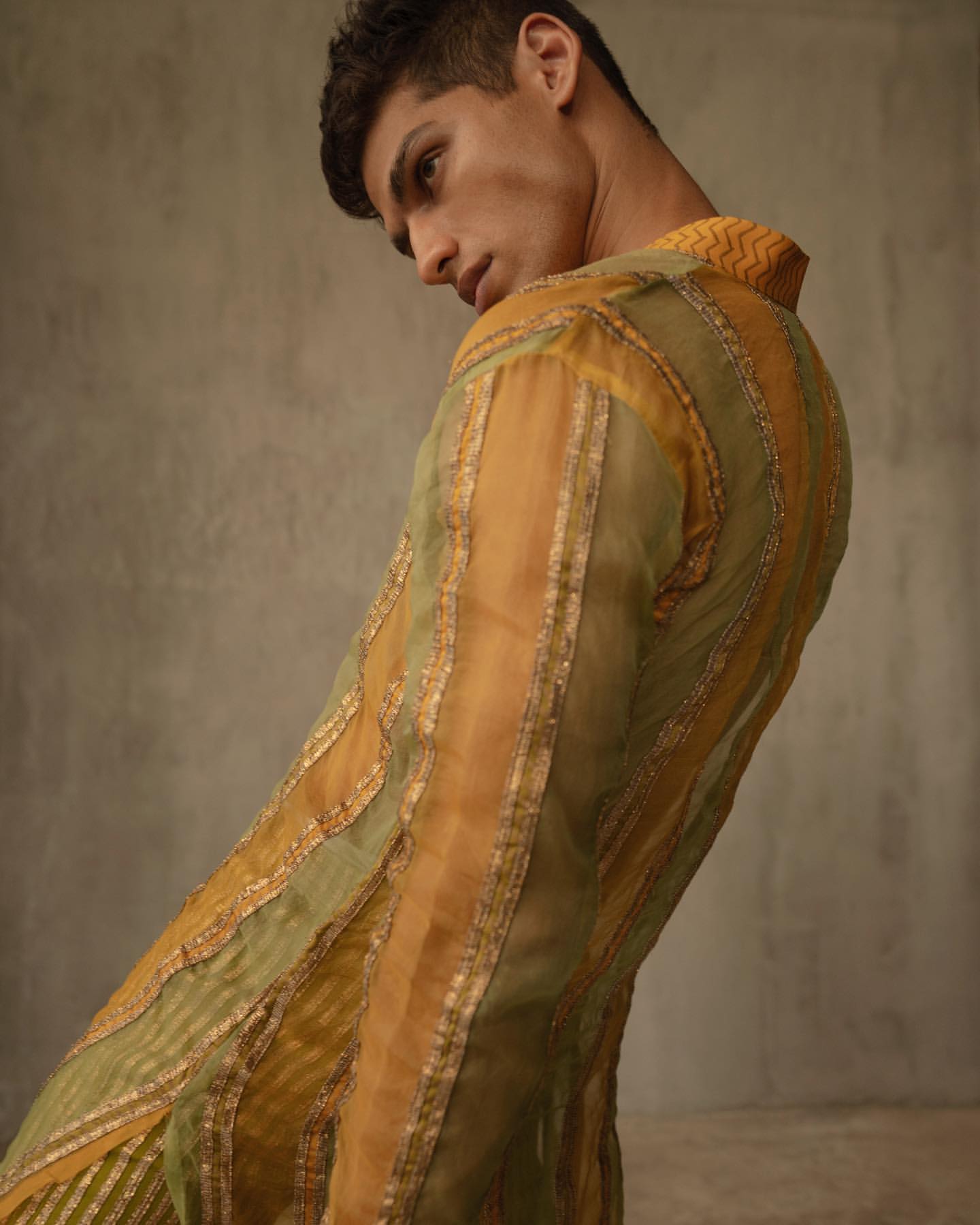 An adult male model poses indoors wearing a yellow and green striped and sheer jacket with a collared shirt and dark hair.