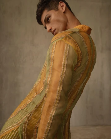 An adult male model poses indoors wearing a yellow and green striped and sheer jacket with a collared shirt and dark hair.