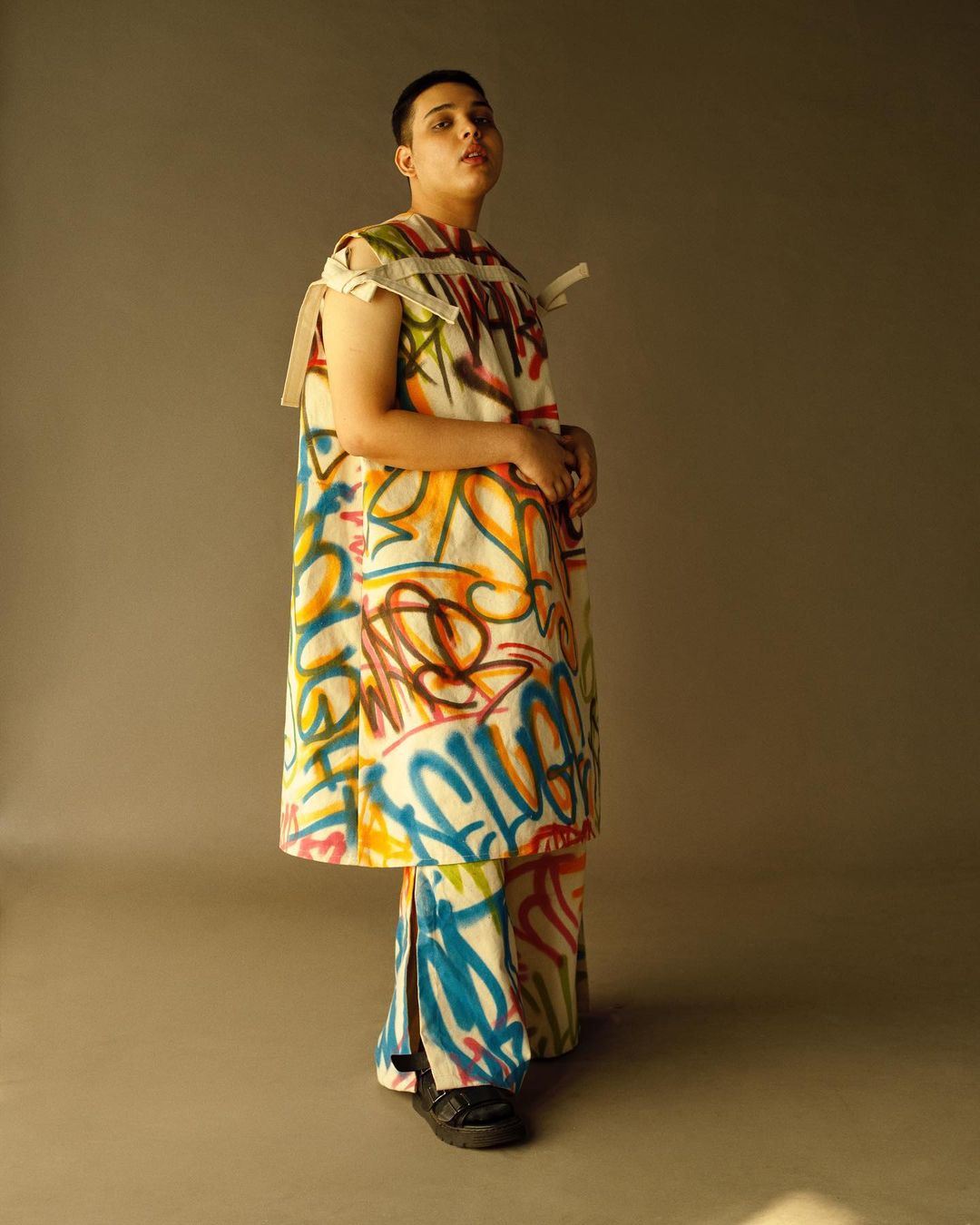 Standing person in a white outfit covered in colorful graffiti-style art, posing against a neutral grey studio backdrop with soft lighting.