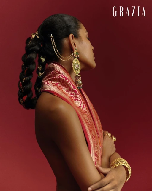 Profile of a woman with a braided updo, draped in a patterned shawl and wearing ornate gold jewelry against a solid red backdrop.