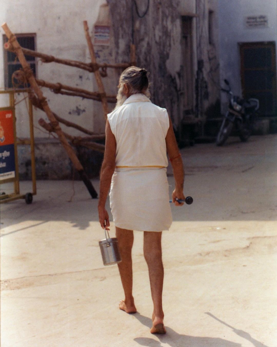 Older man walking in sandals, wearing white sleeveless top and wrap, carrying a small bucket. Background features a sunlit urban area.