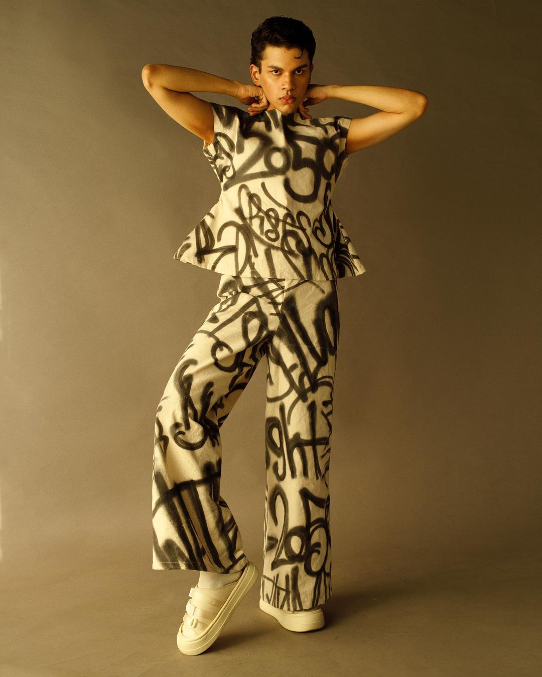 Person wearing graffiti-style top and pants poses in a studio setting with white sneakers. Fashionable pose in coordinated outfit.