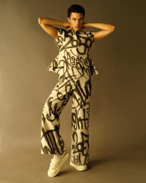 Person wearing graffiti-style top and pants poses in a studio setting with white sneakers. Fashionable pose in coordinated outfit.