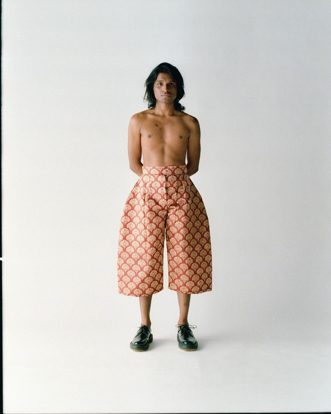 Bare-chested adult male posing in wide, patterned culottes and black dress shoes against a bright white studio backdrop.