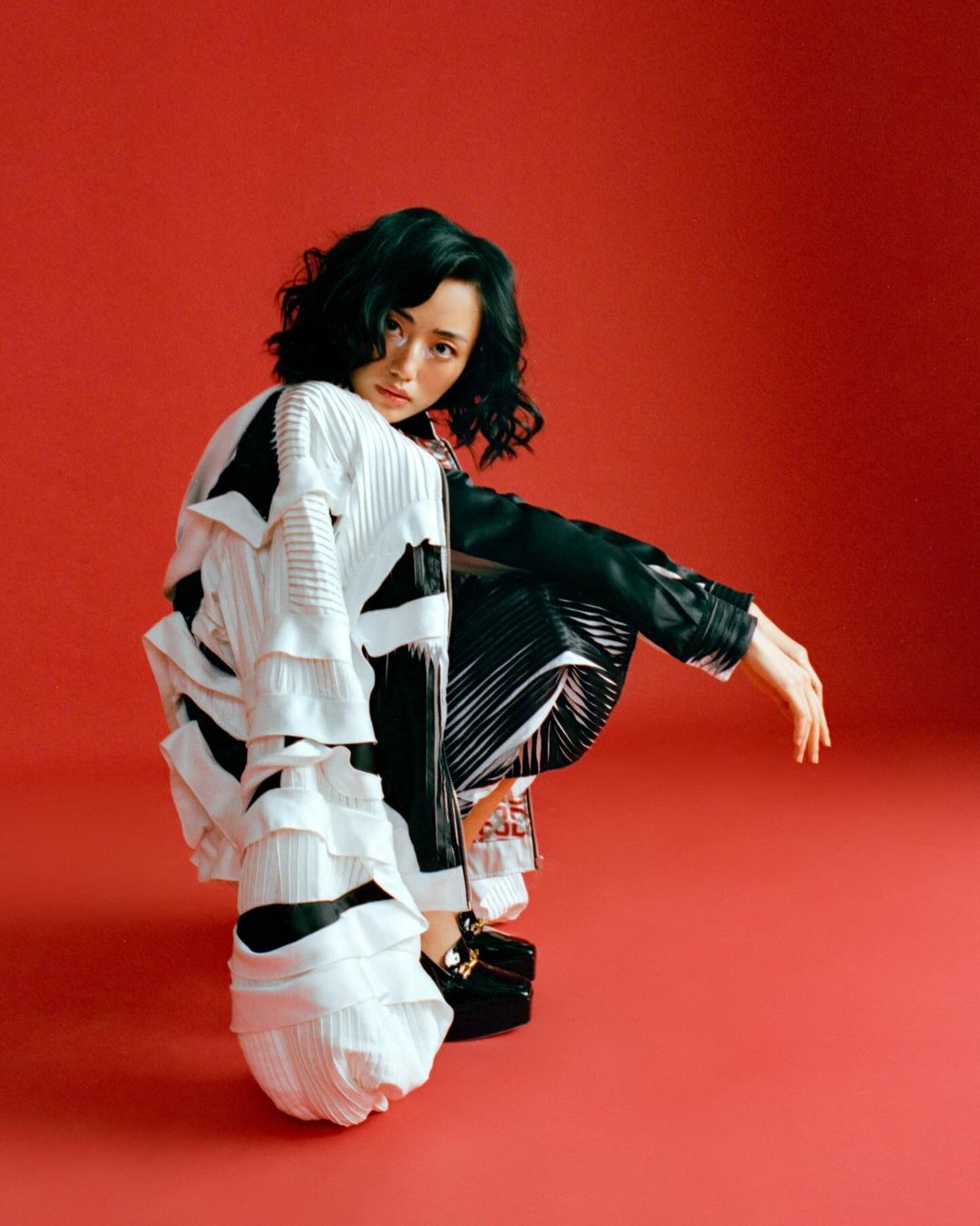 Asian woman in black and white fashion outfit poses in a crouched position against a red background in a studio fashion shot.
