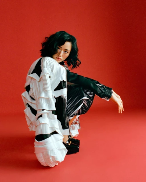 Asian woman in black and white fashion outfit poses in a crouched position against a red background in a studio fashion shot.
