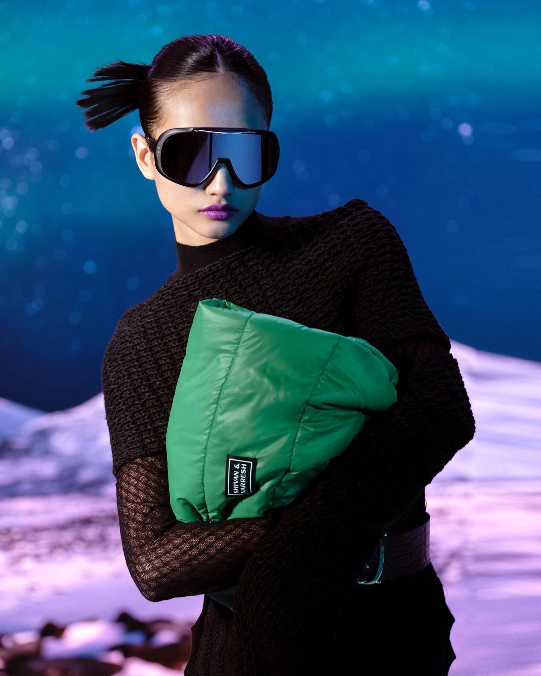 A stylish Asian woman poses in ski goggles, a black turtleneck, and a green bag against a winter mountain backdrop with a blue sky.