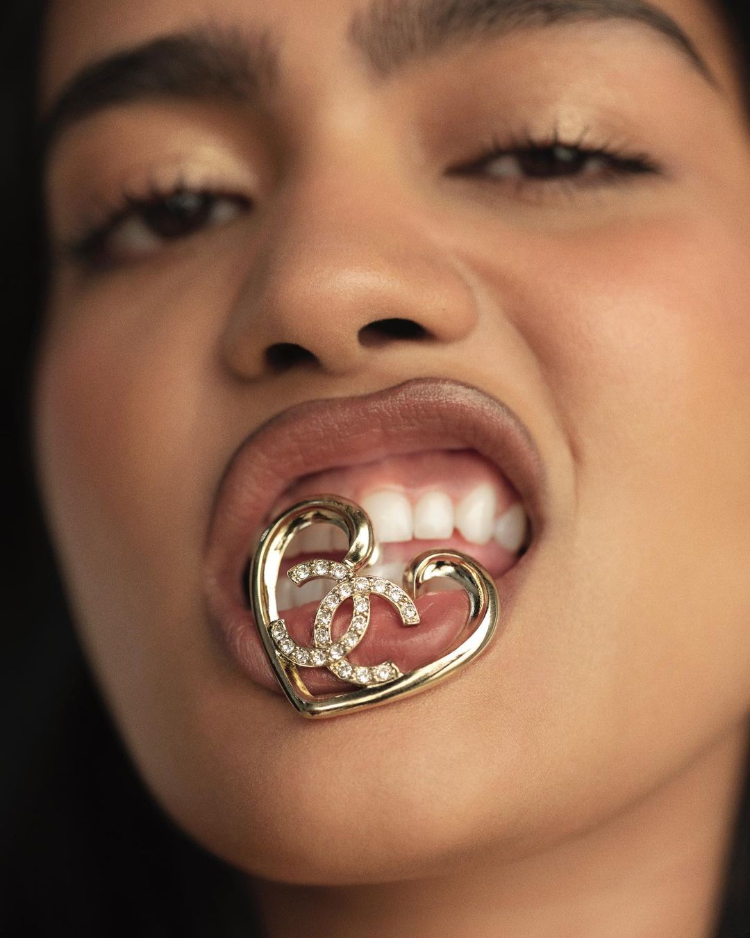 Close-up of a fair-skinned woman holding a gold, heart-shaped Chanel logo between her teeth. Focus is on the jewelry and facial expression.