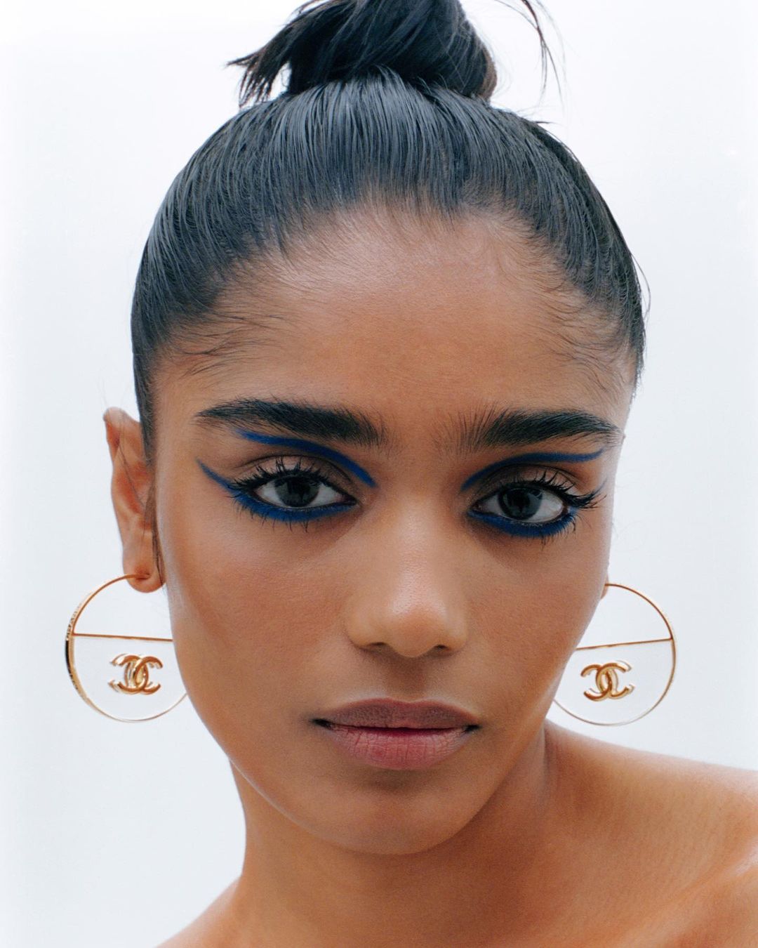 Portrait of a woman with blue eyeliner and hair in a bun wearing hoop earrings with the Chanel logo on a light background.