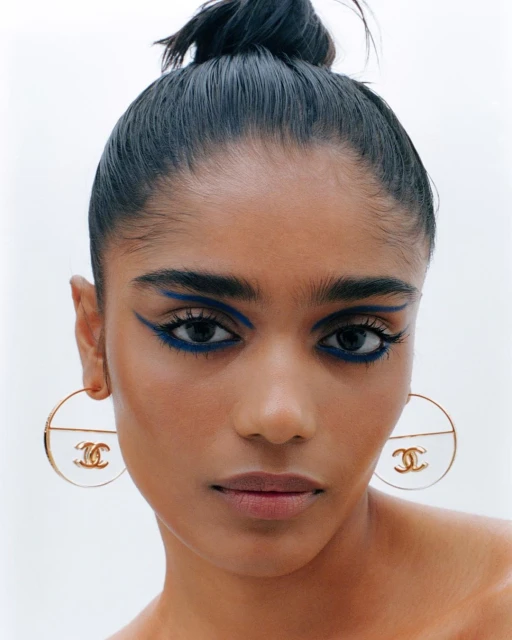 Portrait of a woman with blue eyeliner and hair in a bun wearing hoop earrings with the Chanel logo on a light background.