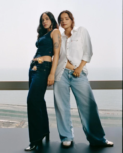 Two young women in denim and white shirts pose indoors with a city view, highlighting contemporary fashion and casual style.