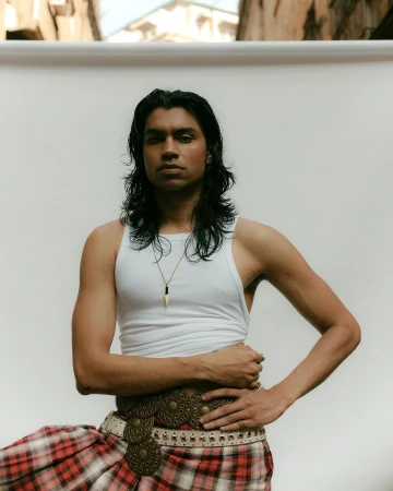 Man with long dark hair wearing a white tank top and plaid kilt against a white backdrop