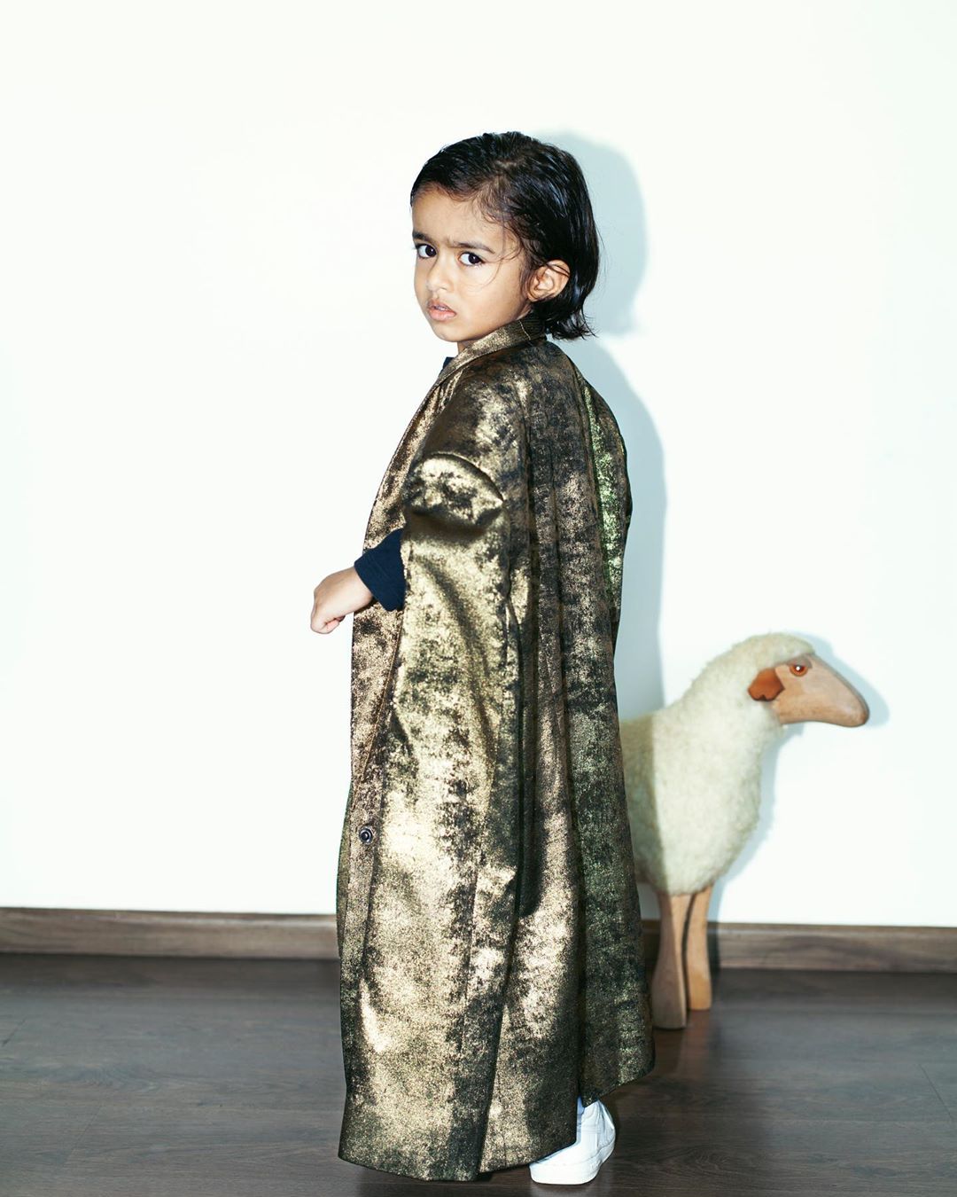 Toddler in a gold cape stands next to a toy sheep in front of a white wall and brown floor, looking back over their shoulder.