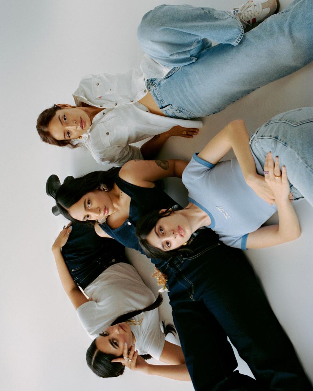 Four women laying on a white surface, wearing different denim washes and tops, in an overhead shot in a fashion editorial.