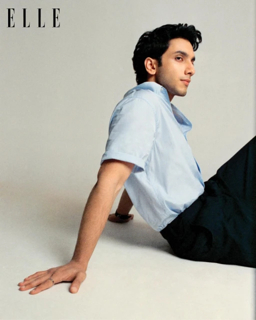 Portrait of a young adult male in a light blue button-down shirt and dark pants, sitting on the floor with a neutral facial expression.