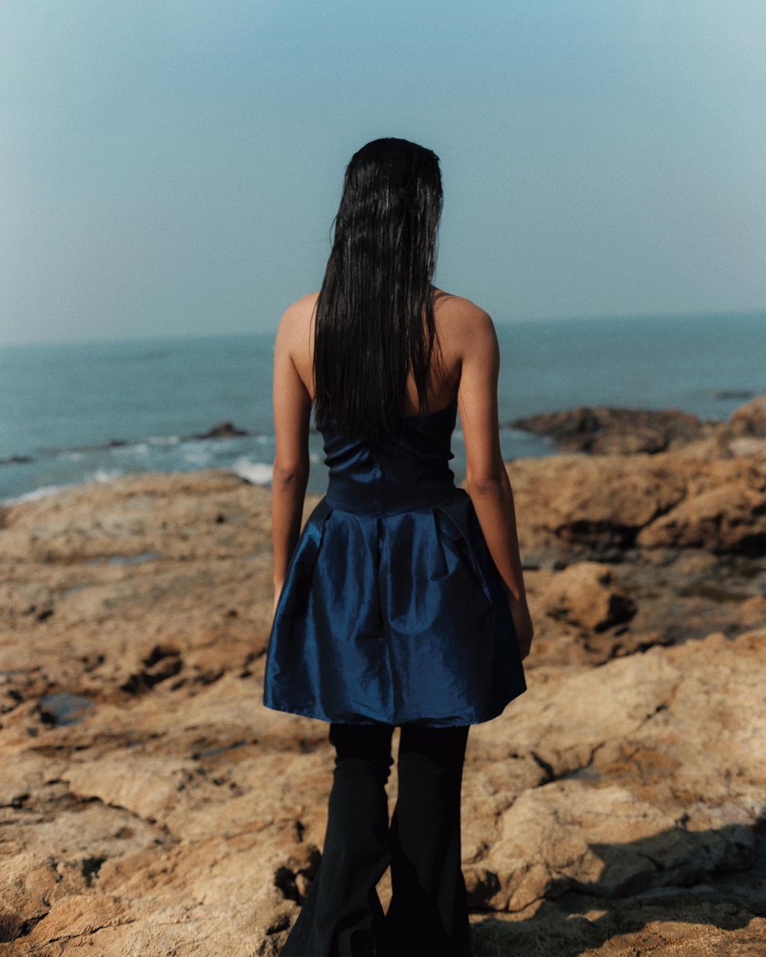 Woman stands on rocky outcrop by the sea. She is wearing a strapless blue dress and black leggings, facing away from the camera.