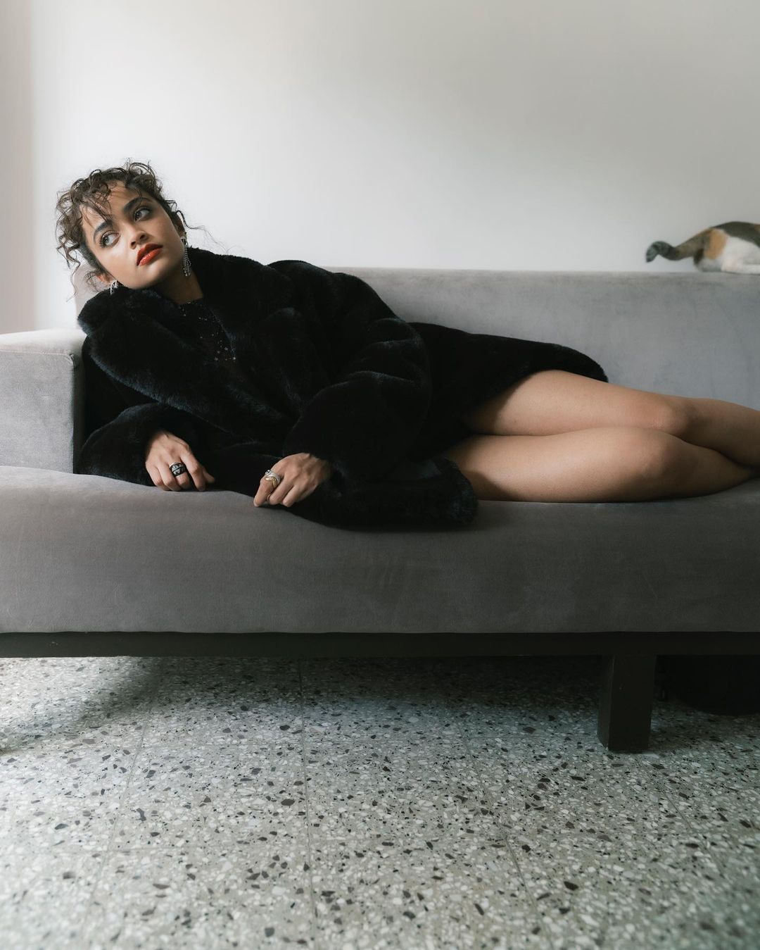 Young woman in a black fur coat rests on a gray couch in an indoor setting, accentuated by cool, natural light and a terrazzo floor.