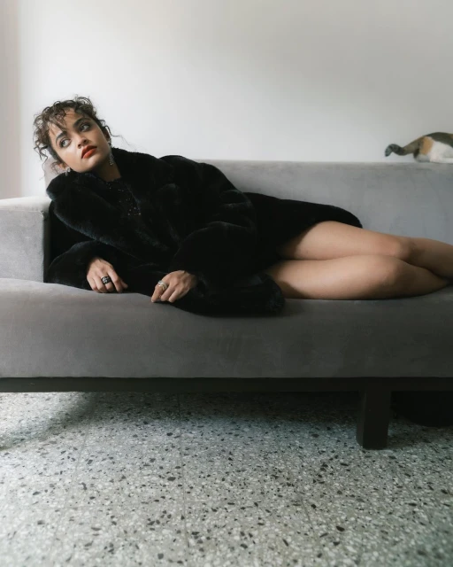 Young woman in a black fur coat rests on a gray couch in an indoor setting, accentuated by cool, natural light and a terrazzo floor.